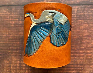 Features a flying blue heron hand tooled and painted design on a saddle tan brown napkin ring. On a timber background.