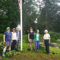 A group of garden club members stands proudly in front of the Hanson Family Memorial garden, with the American flag raised behind them. They are dressed in gardening attire, including hats, gloves, and boots, and smile warmly at the camera. The garden they have tended to features a variety of lush plants and flowers, creating a peaceful and vibrant atmosphere around the memorial plaque. The tall trees and greenery in the background add to the serene setting.