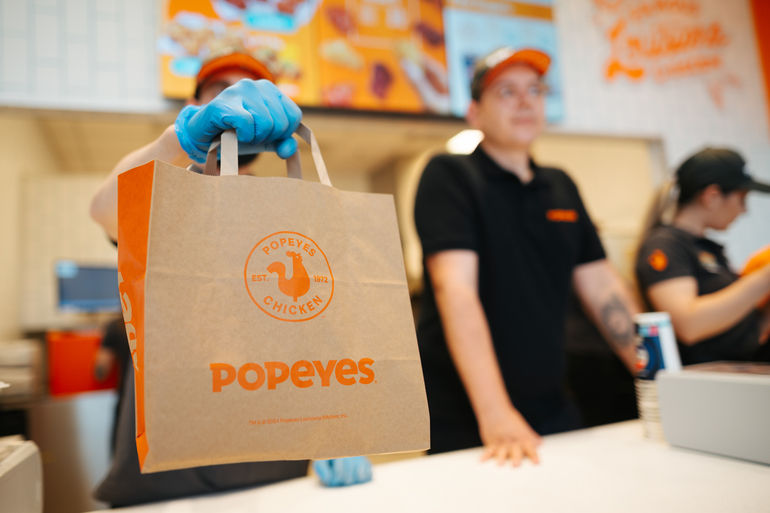 Angajat Popeyes cu mănuși albastre ține o pungă maro inscripționată. Fundal cu meniuri colorate, angajați în uniformă neagră. Atmosferă activă.