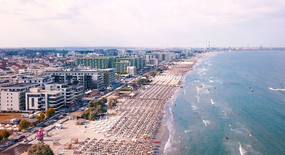 Plajă aglomerată lângă oraș, umbrele albe, clădiri moderne în plan apropiat, valuri calme, cer senin, atmosferă relaxantă.