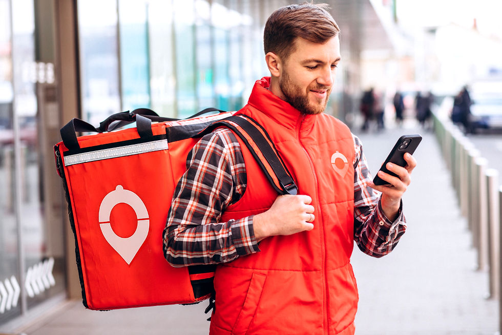 Curier îmbrăcat în vestă roșie, cu un rucsac mare, se uită la telefon pe stradă. Fundal urban, vreme însorită. Echipament portocaliu.