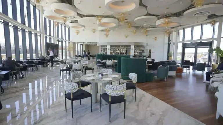 Restaurant modern cu podea de marmură, scaune cu modele alb-negru, tavan elegant, lumini rotunde. Barul în fundal, ferestre mari. Atmosferă elegantă.