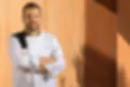 Bărbat în uniformă de bucătar, cu brațele încrucișate, zâmbește. Fundal din lemn. Pe haină scrie "PALMARES" și un nume. Lumina caldă.