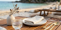 Picnic table with plate, glass, and gold cutlery on sandy beach.