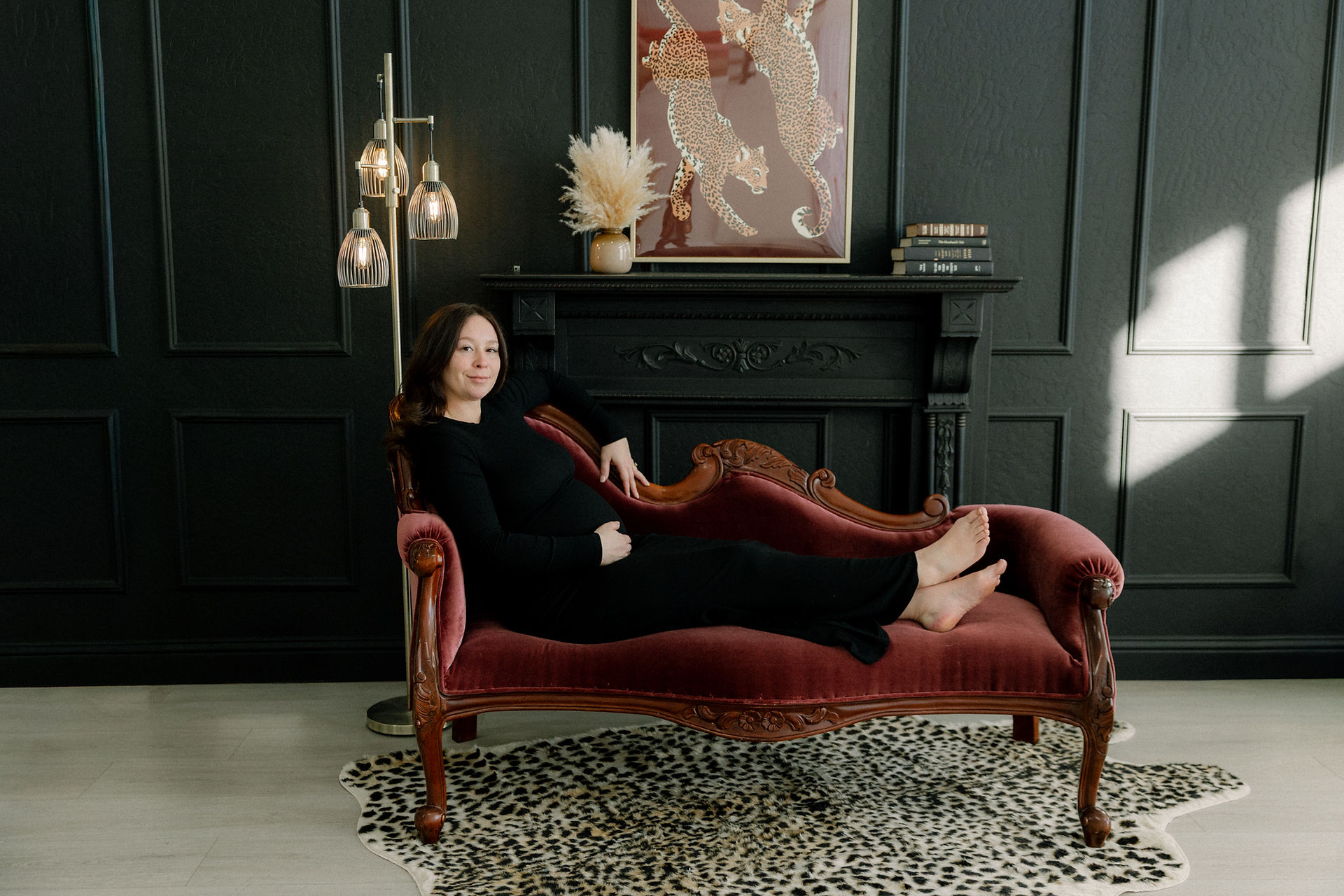Maternity photo of mom-to-be in black dress sitting on maroon chaise lounge