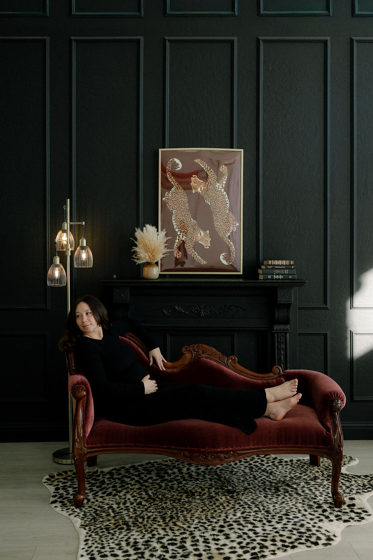 Maternity photo of mom-to-be in black dress sitting on maroon chaise lounge