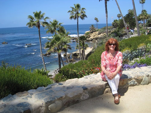 Woman in a pink shirt sits on a stone wall by the ocean, surrounded by palm trees and greenery. Clear blue sky and sea in the background.