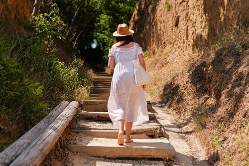 Barefoot woman in a white dress and hat walks up stone steps in a sunlit, sandy path surrounded by greenery. Calm, summer setting.