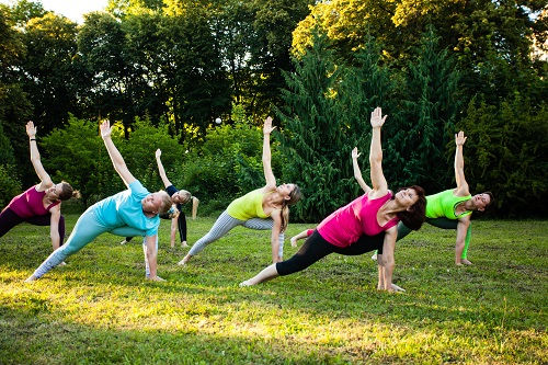People practicing yoga outdoors on grass. They are in side plank poses, wearing colorful attire. Trees in the background, sunny setting.