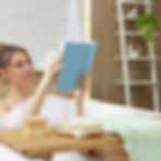 Woman in a bubble bath reads a blue book, surrounded by candles. Wooden tray holds soap and brush. Ladder shelf in background. Relaxing mood.