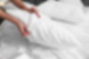 Hands adjusting a white pillow on a neatly made bed with white linens. Soft lighting creates a calm, tidy bedroom atmosphere.