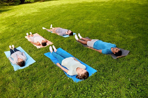 Five people lying on yoga mats in a green park, eyes closed, practicing relaxation or meditation. Bright, sunny day.