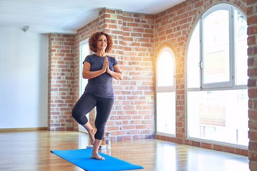 Woman practicing yoga in a bright room, balancing on one leg in tree pose on a blue mat. Brick walls and arched windows in the background.