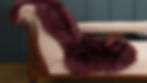 A plush maroon sheepskin rug drapes over a beige chaise lounge with carved wood accents. An open book lies on the tan carpet. Dark paneled wall.