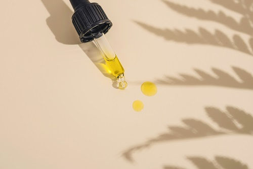Dropper with yellow oil spills onto beige surface, next to plant shadows. Minimalist and calm mood, no text present.