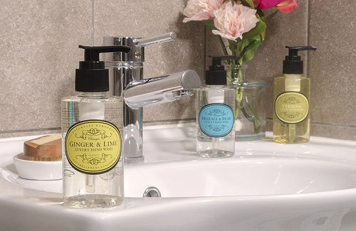 Three hand soap bottles on a bathroom sink beside a chrome faucet, with flowers in a vase and a calm, clean feel