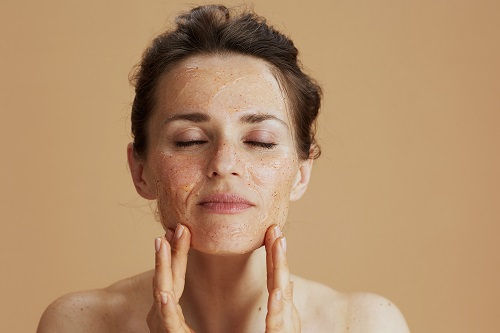 Woman with closed eyes applying facial scrub, beige background. Her expression is relaxed and serene.