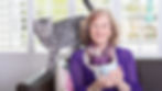 Woman in a purple blouse sits on sofa with a mug, alongside a striped cat. Bright window shutters and floral cushion in the background.