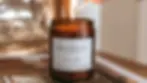 Hand lighting an amber candle labeled "Geodesis Figuier" with a match. Candle rests on a magazine, with a blurred background. Cosy mood.