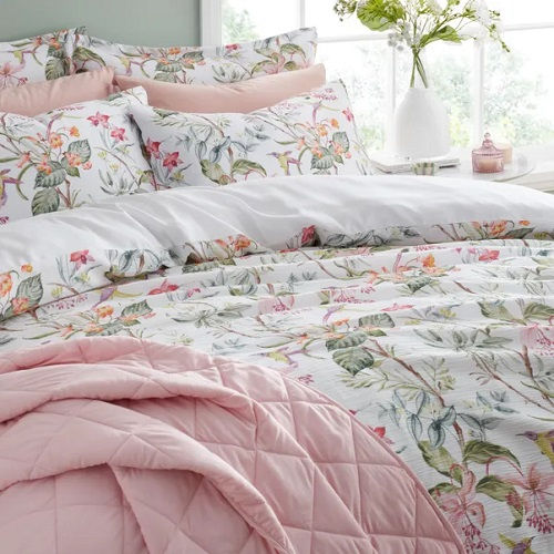 Floral-patterned bedding with pink quilt, set in a bright room. A vase with flowers on the window sill adds a fresh, serene mood.