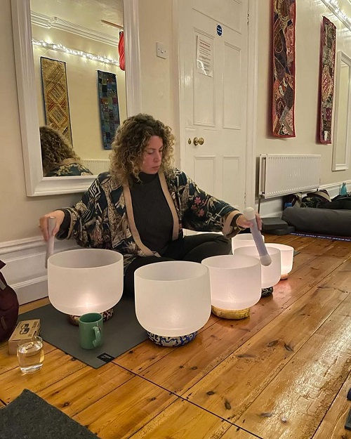 Person plays crystal singing bowls in a serene room with wooden floors and decorative wall hangings. Warm lighting creates a calm atmosphere.