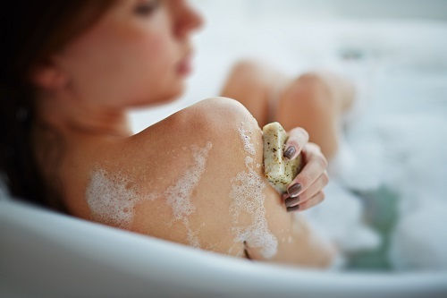A woman in a bathtub scrubs her soapy back with a soap bar, relaxed and blurred in a bright bathroom.