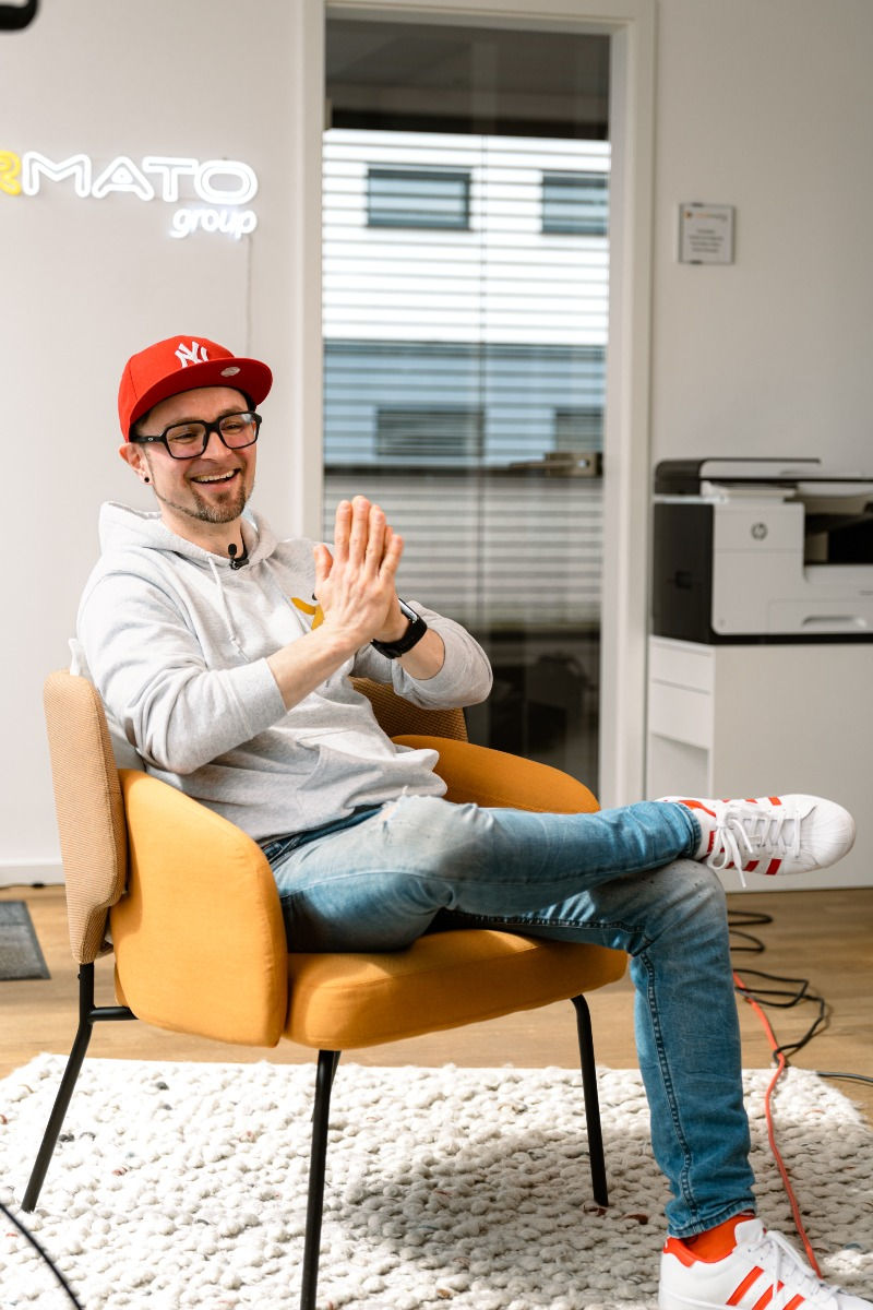 Mann in legerer Kleidung und roter Baseball-Cap lacht sitzend im Stuhl