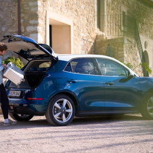 Mann lädt einen silbernen Koffer in den Kofferraum eines blauen Autos.