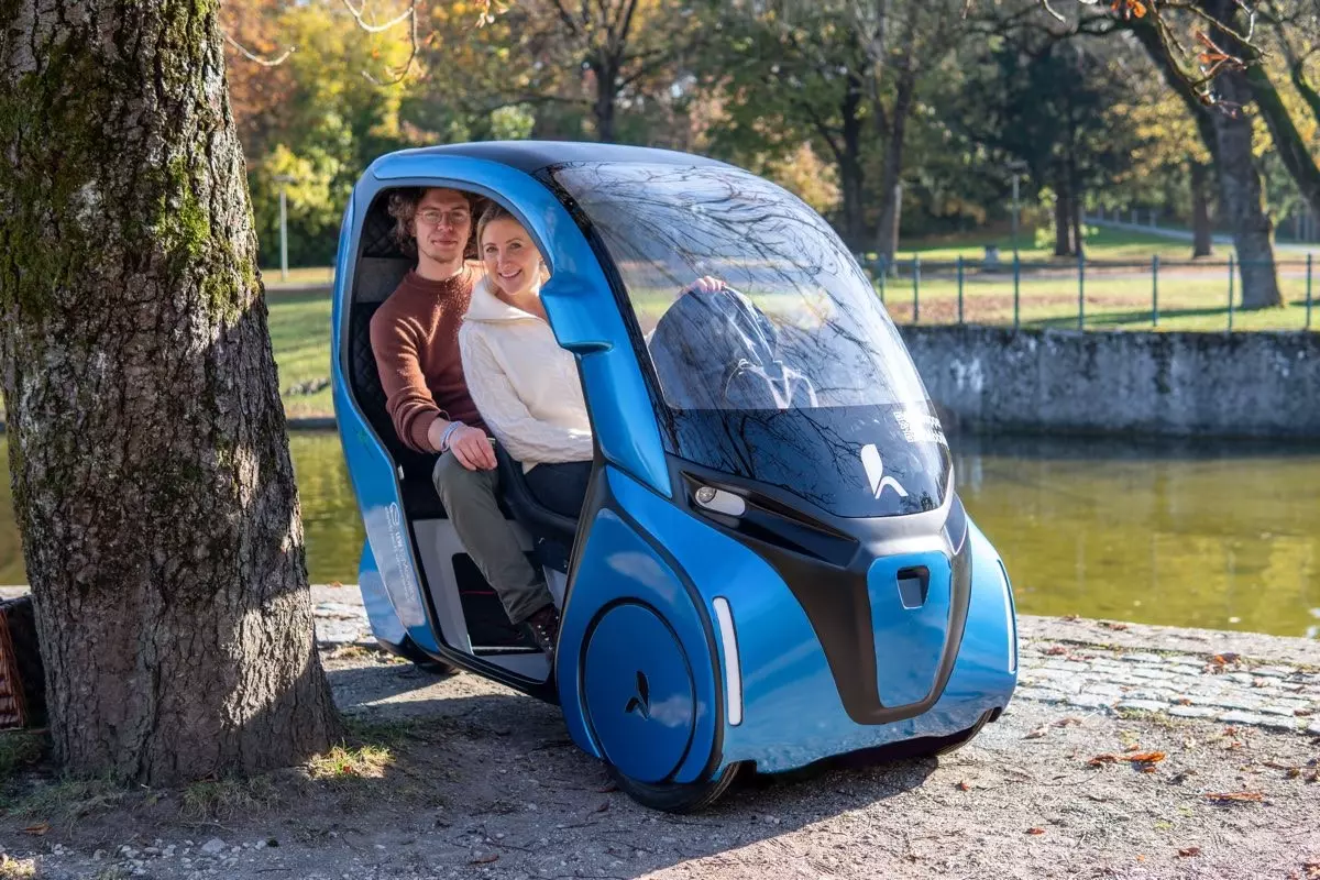 Mann und Frau sitzen in einem Kabinenartigen Auto mit Pedalantrieb.