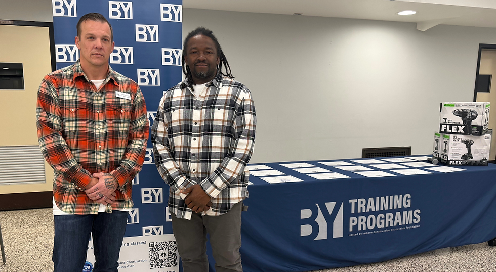 two men stand in front of a BY Plus banner and table.