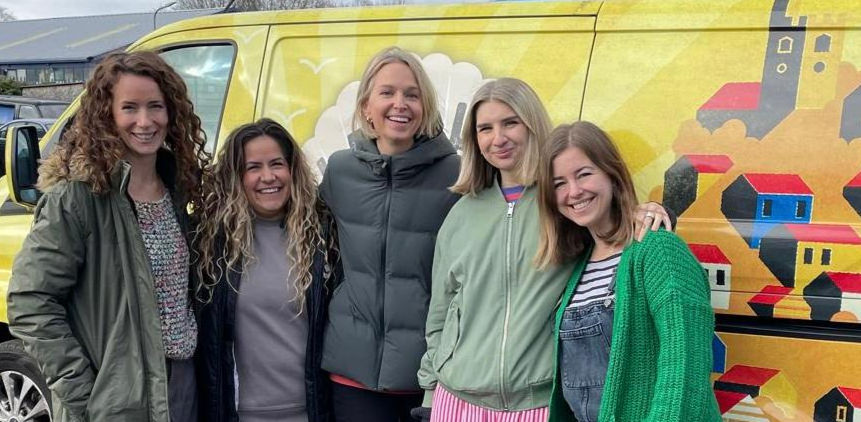 A group of five middle aged women in warm clothes standing next to a yellow van, smiling.