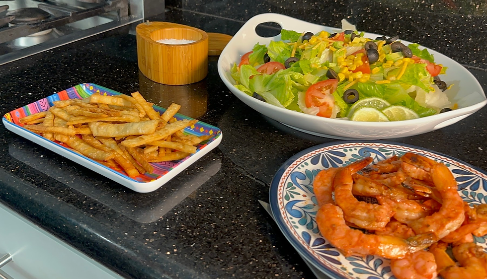 Colorful plates of fried tortilla strips, shrimp, and a fresh salad on a black kitchen countertop with a salt container nearby