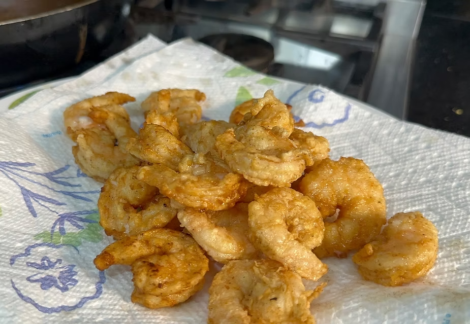 Pile of golden fried shrimp on a floral paper towel in a kitchen, glistening and freshly cooked on the stovetop.