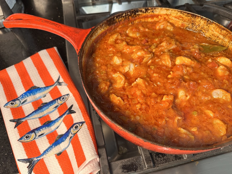 Red skillet on a stovetop simmers orange stew, beside a striped towel with blue fish in a cozy kitchen setting.