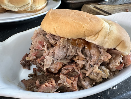 Close-up of a pulled pork sandwich on a white plate, with shredded meat spilling from a soft bun on a kitchen counter.
