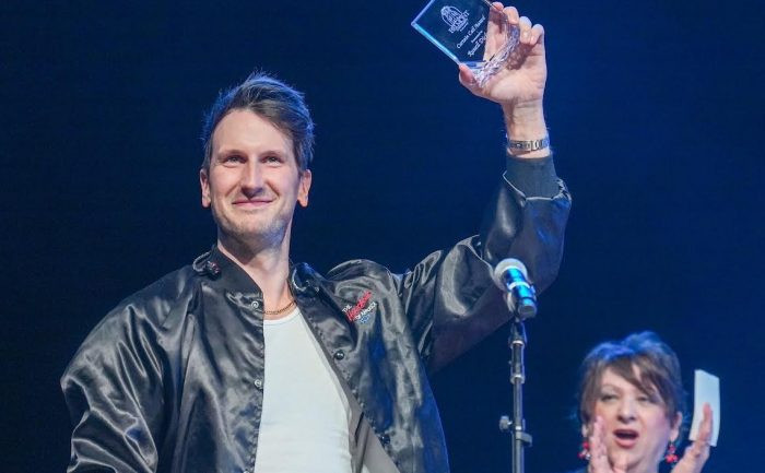 Russell Dickerson holding up a glass award while Sandra Dudley applauds behind him