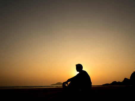 Silhouette of a person sitting on a beach at sunset, with orange sky, calm sea, and distant cliffs.