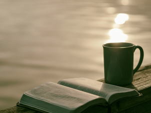 Open book and green mug on a wooden railing by a shimmering lake at sunset, creating a serene and reflective mood.