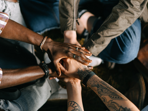 Hands stacked in a group, displaying unity and diverse tattoos. Outdoors on grass, casual attire visible, conveying a sense of teamwork.