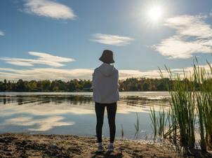 Person in a hoodie and hat stands by a lake, facing the sun. Tall grass and sandy shore in the foreground, trees in the background. Peaceful scene.