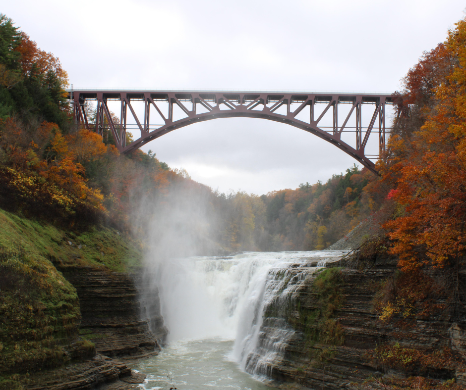 Waterfall flows beneath a tall iron bridge, surrounded by vibrant autumn trees. Mist rises, creating a serene, natural landscape.