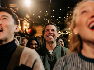 A joyful crowd of adults of different ages under string lights, smiling and laughing together in a dimly lit venue like a comedy show. Warm, festive atmosphere with a focus on positive emotions.