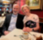A photo of Mary, the subject of this article, and her husband Pat, smiling and seated at a restaurant table with coffee and cake, in front of red walls and a gilded painting.