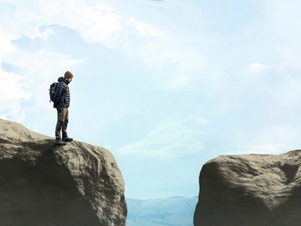 Man stands on a cliff edge, gazing at a gap. Clear sky and distant mountains in the background, conveying contemplation.