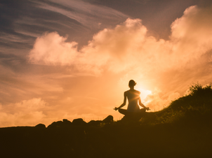 A person sitting criss-cross with their arms open meditating on a mountain with the sunrise in the background coming through the clouds