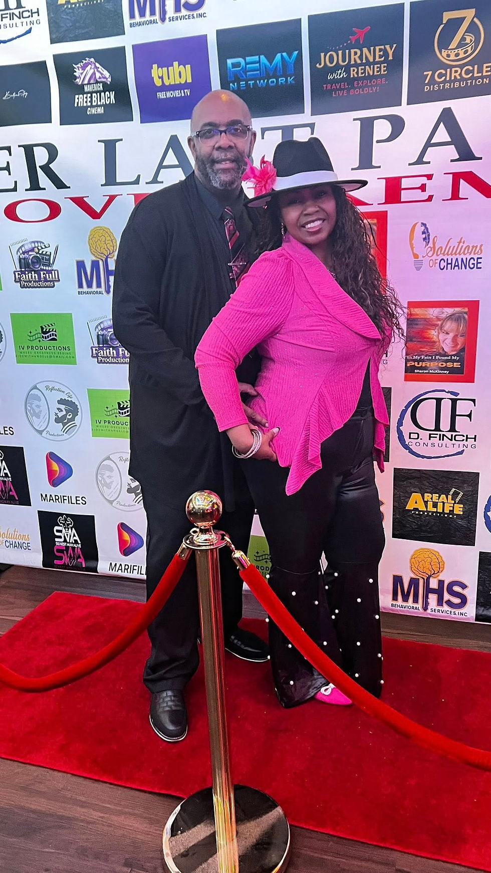 A man in a black suit and a woman in a pink outfit with a hat pose happily on a red carpet. Logos and event text in the backdrop.