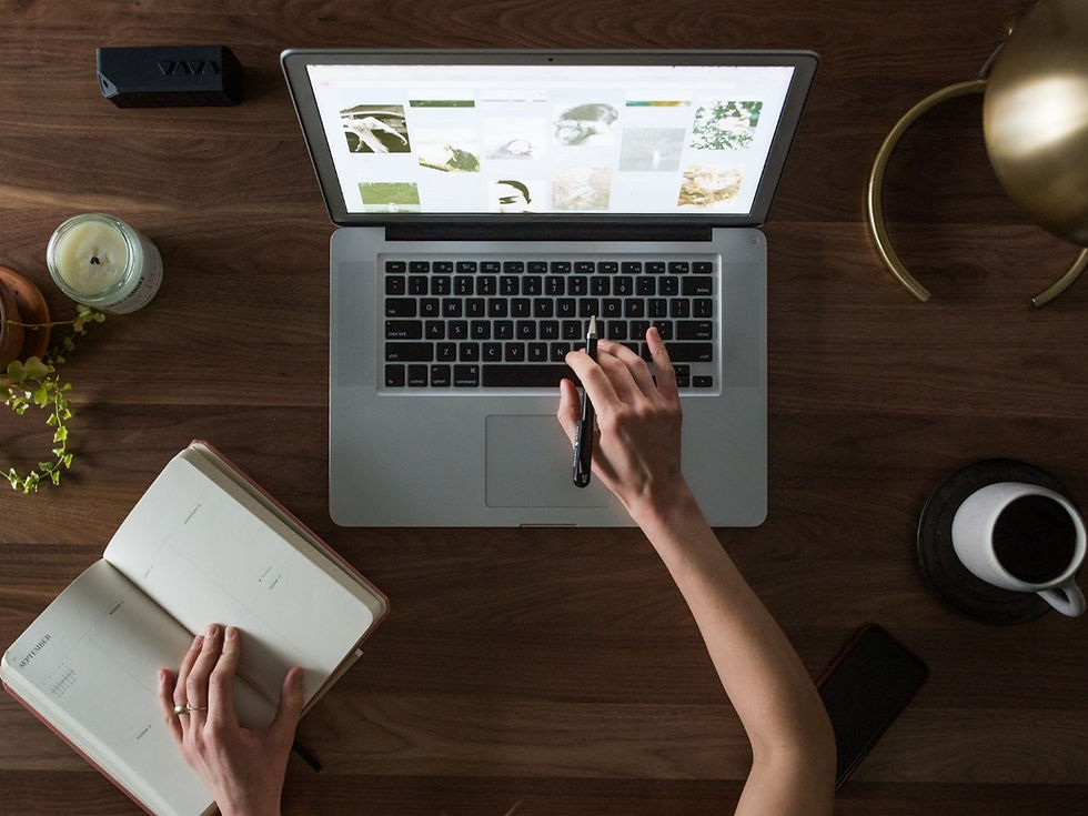 Person using a laptop with images, holding a notebook and pen on a wooden desk. A candle, plant, and coffee cup are nearby. Cozy setting.