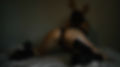 A woman in black lingerie and platform heels poses on a bed, facing the wall with her hands raised and back arched. The lighting is moody and intimate, emphasizing the curves of her body and the softness of the setting.