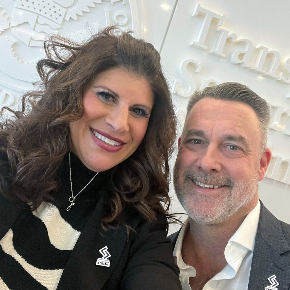 Two people smiling indoors, wearing SYNect logos. Background shows embossed text and an eagle seal on a white wall, creating a professional tone.