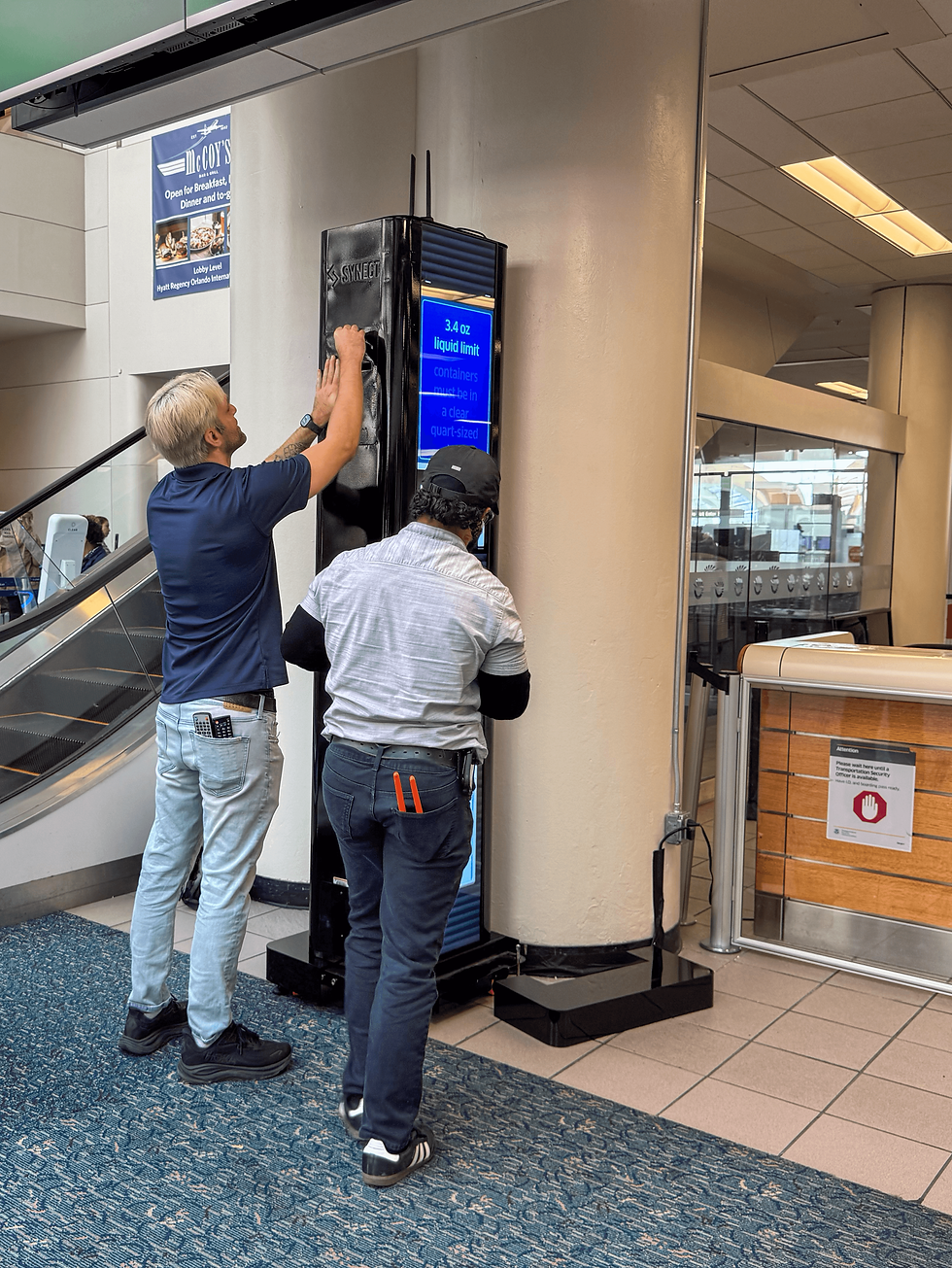 Two men install a black digital sign in an airport lobby; blue screen shows 3.4 oz liquid limit, near stairs and glass doors.